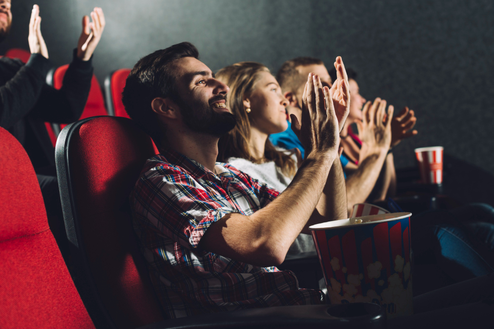 Pessoas no cinema com balde de pipoca no colo e aplaudindo.